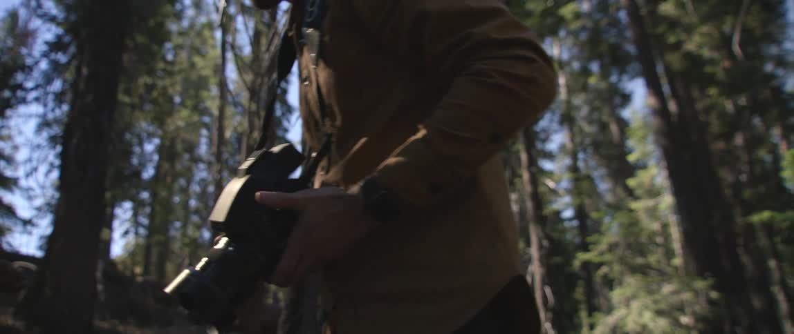 Male photographer with brown shirt and black pants carries camera in the forest