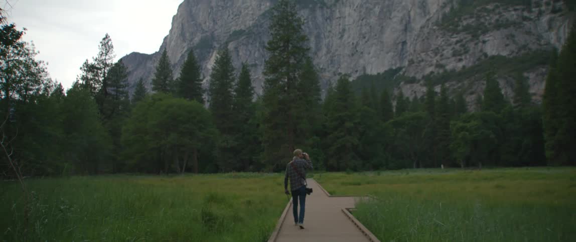 Man with a camera walks past a field and towards a forest and the mountains