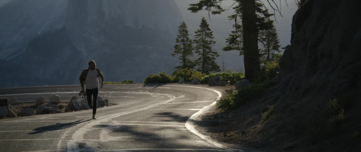 A man holding a skateboard running on a road with a mountain range the sky and clouds in the background