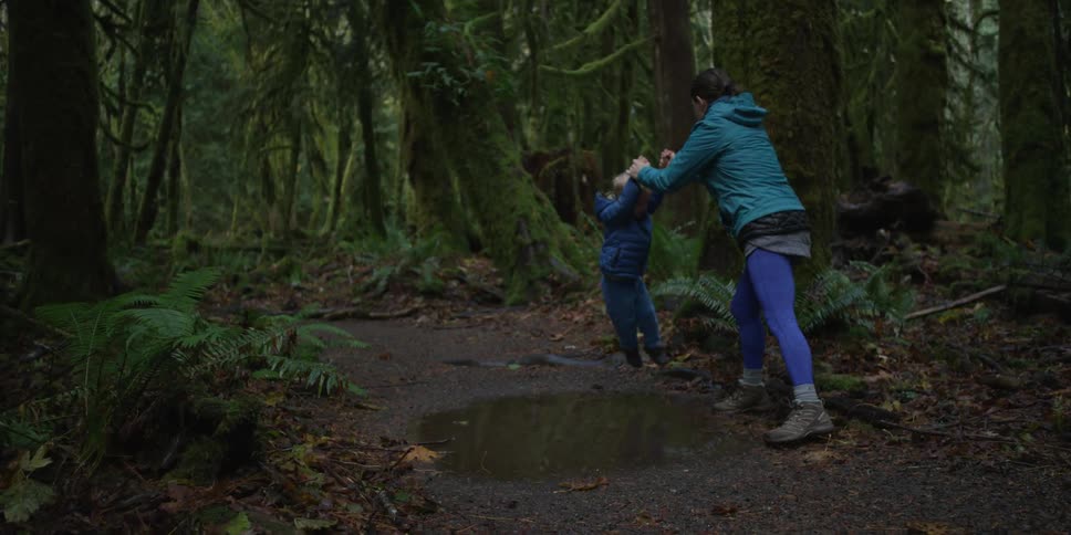 A woman lifts a boy over a pond in the woods