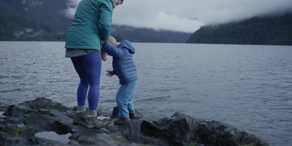 A woman holds the hand of a toddler as they balance on rocks near a shore