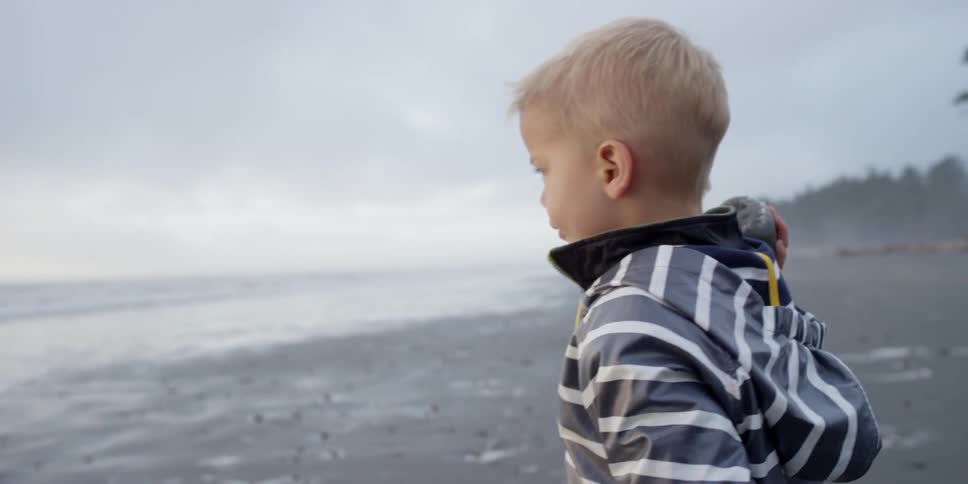 A boy standing on a beach holds a rock over his shoulder