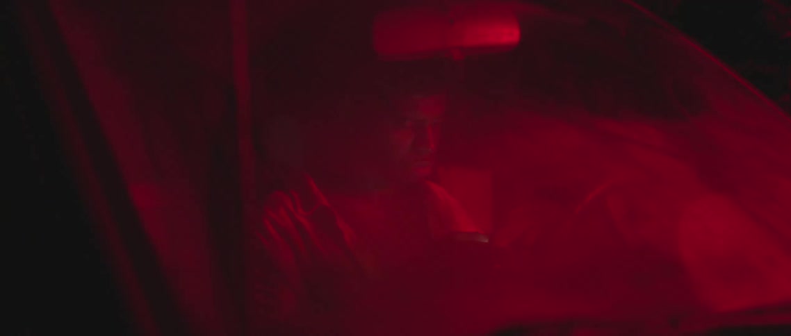 Young man looking at his phone sitting on a car under red lights at night