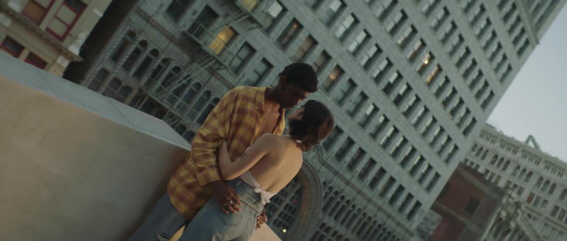 A young man and woman stand close and embrace atop a building in a metropolitan area