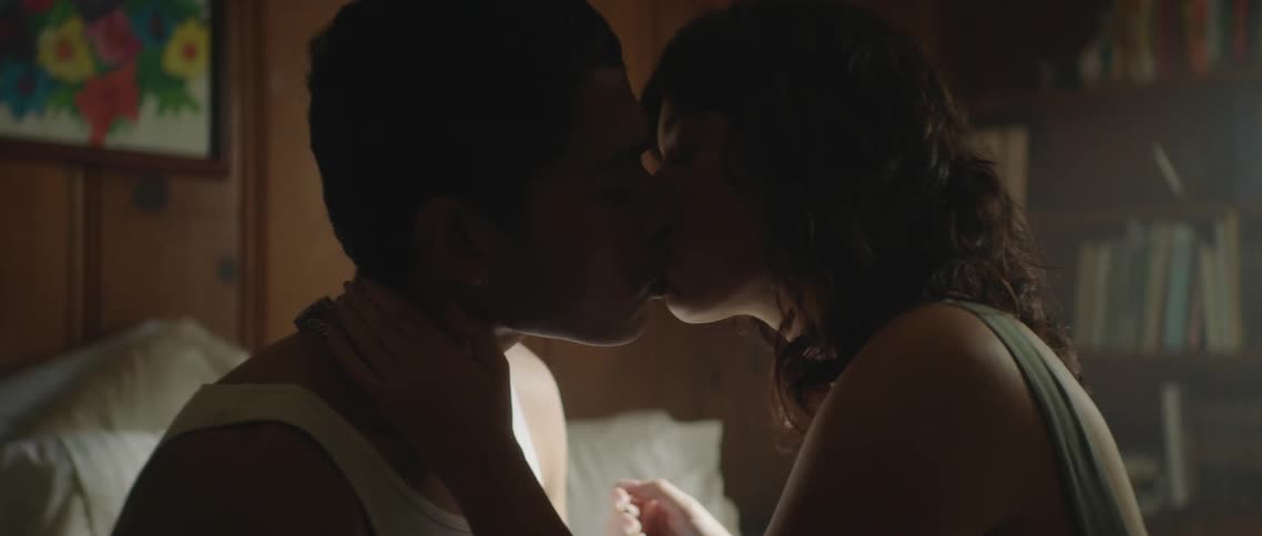 A young man and woman sit on the bed together while lovingly kissing each other