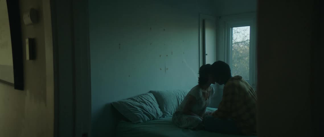 A young couple lean forward to kiss each other while sitting on a bed in a dimly lit room