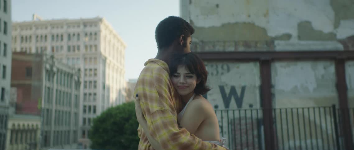 A teenage girl and boy stand in front of a building in a city and hug one another