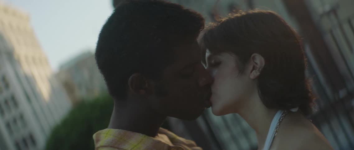 A man and a woman on a rooftop kissing with buildings the sky and clouds in the background