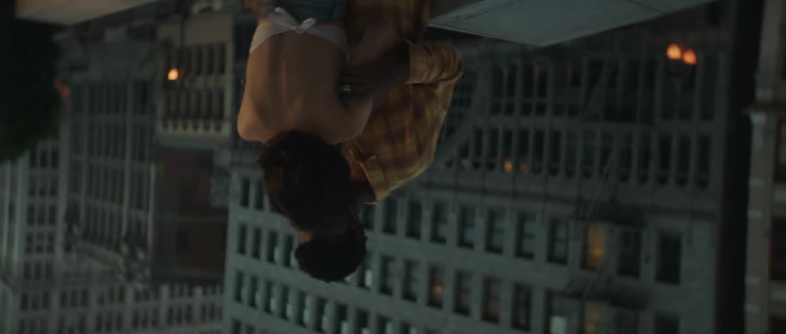 The camera spirals away from a young man and woman as they embrace on the rooftop of a tall building