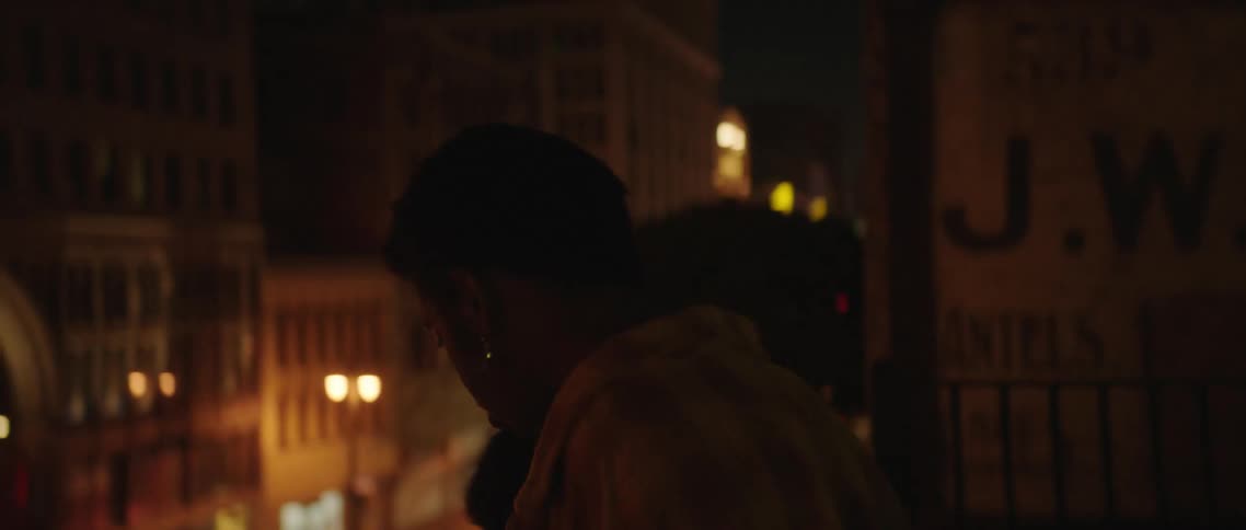 Man and woman stand on rooftop at night looking around city street