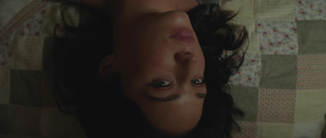 A woman lying atop a bed turns her head to look up into the camera