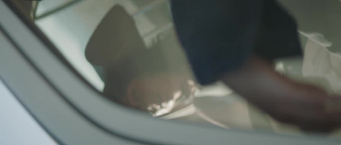 A young man and woman sit in a car as the camera spirals just outside of the driver side window