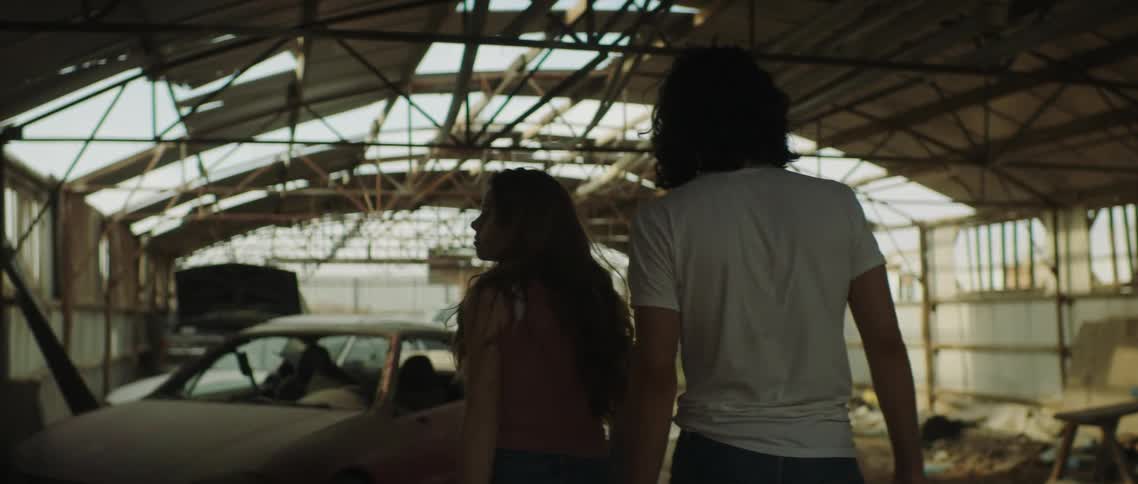 A man and a woman walking in an abandoned hanger with vehicles with the sky and clouds in the background