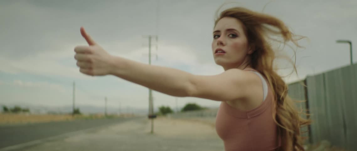 A young woman with long hair defiantly walks away from her male companion while she holds out her thumb to hitchhike