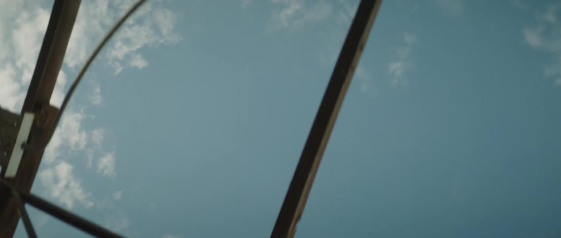 A young couple stand close under the open frame of a building as the camera pans upwards at the sky above them