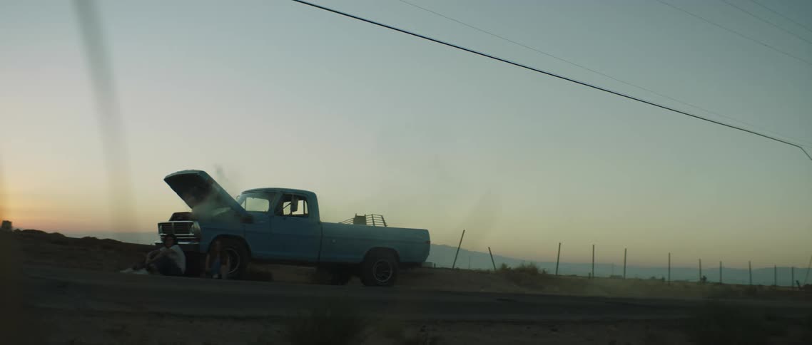 Two people are sitting next to a car which seems to be damaged as smoke seems to be coming from the engine