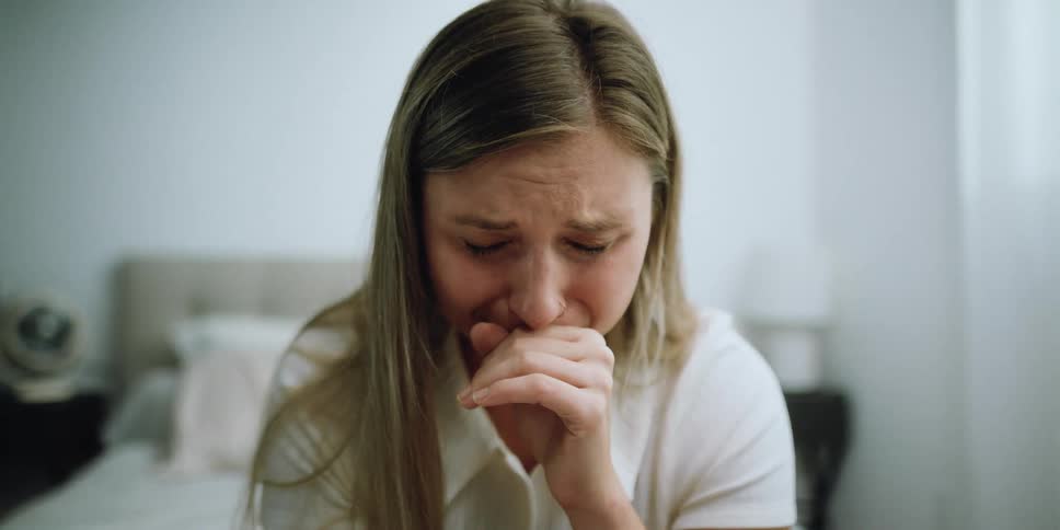A woman in a room sitting on a bed crying with a wall and window in the background