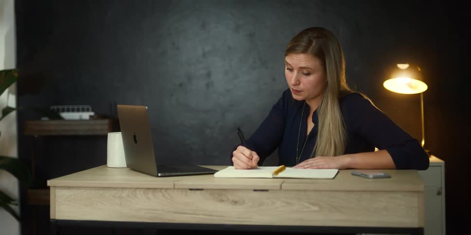 A woman is taking notes while watching a laptop at work