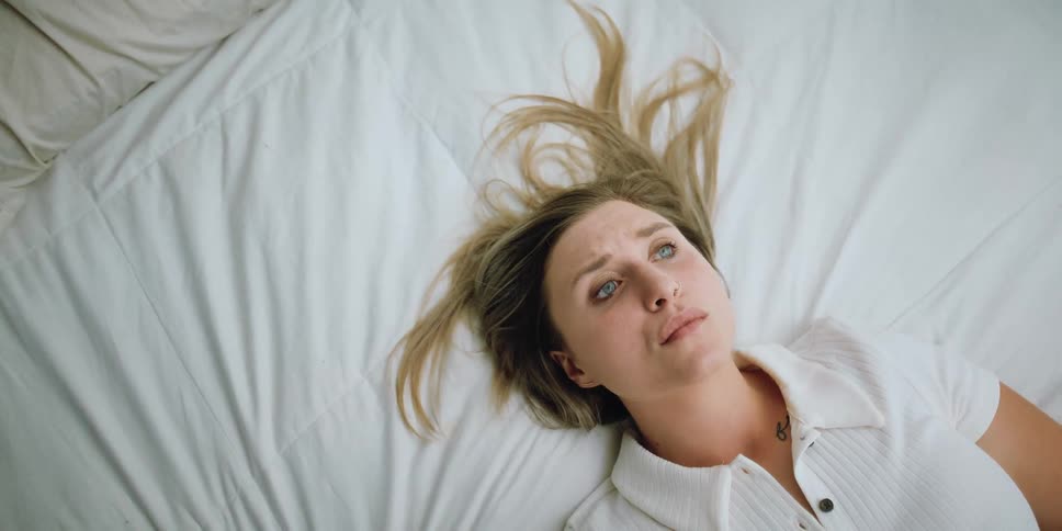 Blonde woman with white shirt lies down on bed and is being emotional after looking at baby photos