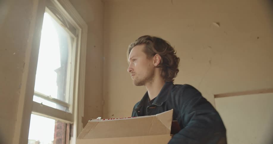 A man holds a cardboard box and looks out the window of a run down apartment