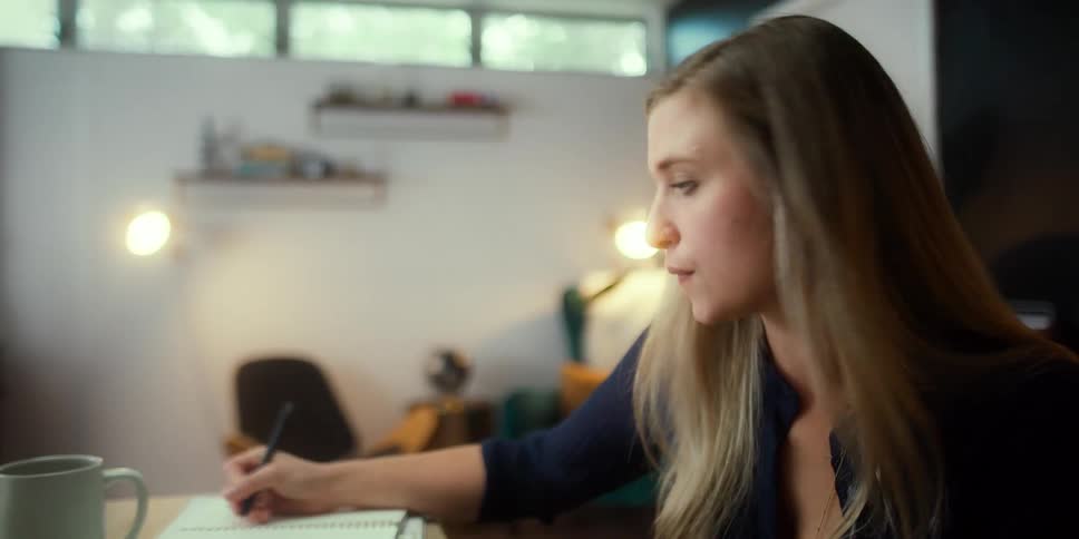 A woman in a room writing on a notebook with a pen and looking at a laptop with a wall in the background