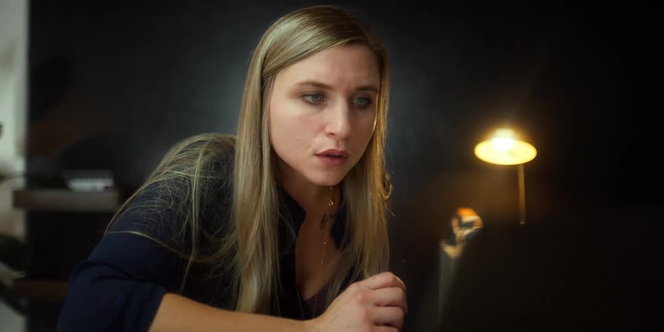 Woman with long blonde hair watches something on her laptop as a lamp sits on a dresser behind her
