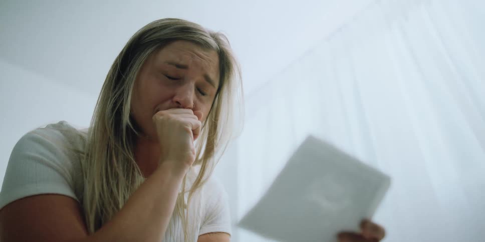 A woman squeezes her eyes shut while crying as she looks at the page she is holding