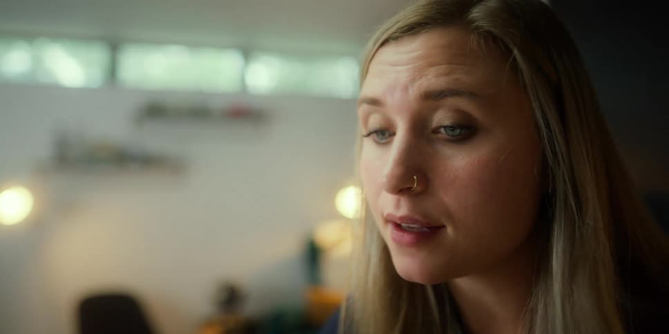 A woman with a pen in a room talking with a wall and window in the background