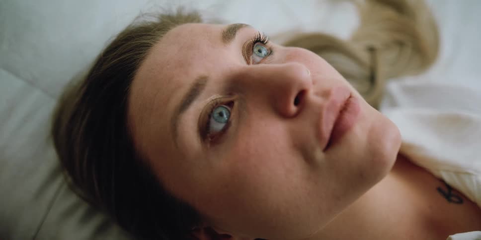 Blonde woman with blue eyes and white shirt lies down on bed pondering
