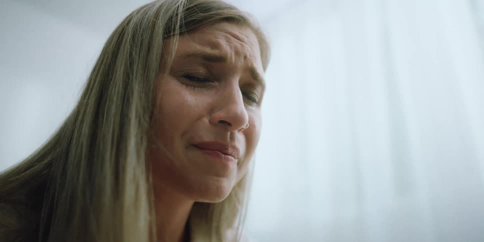 A woman in a room crying with tears in her eyes with a wall in the background