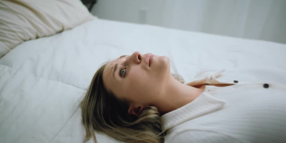 Blonde woman with white shirt lies down on bed looking sad in bedroom