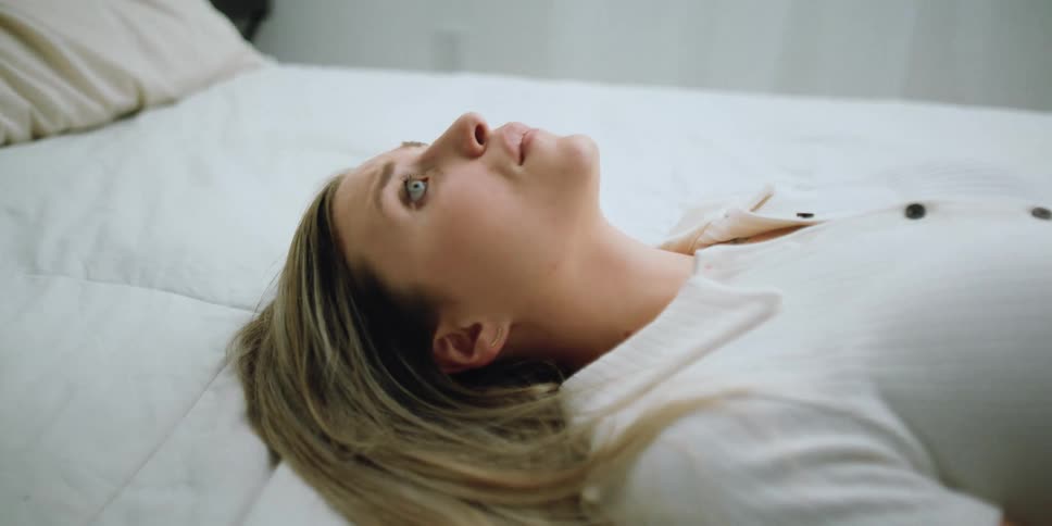 Blonde woman with white shirt and pants lies down on bed looking upset