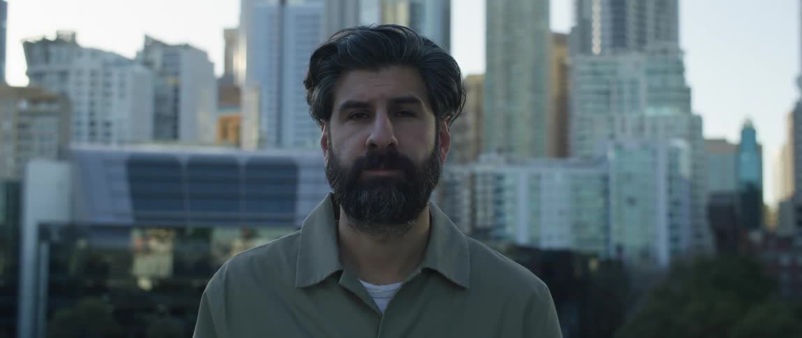 Male photographer with beard and shirt looks at camera with high rise buildings in the background