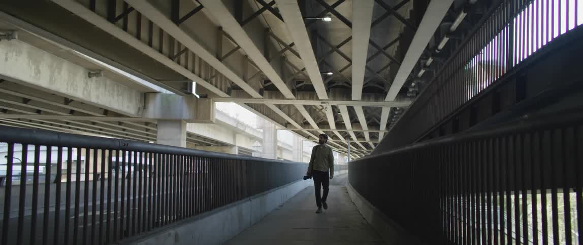 A man with a camera walking underneath an overpass with the sky and clouds in the background