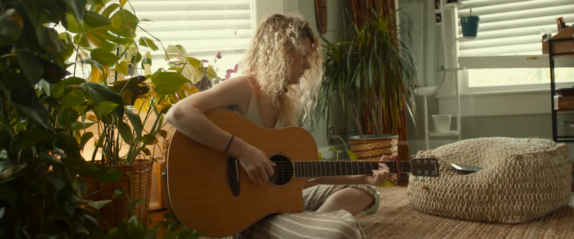 Woman sits cross legged on rug playing acoustic guitar near houseplants and windows