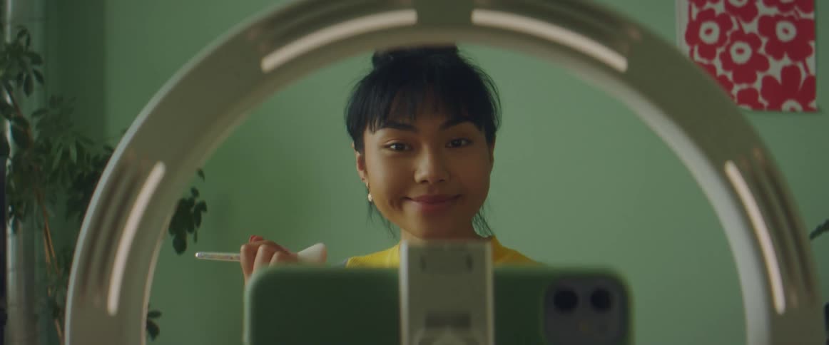 Woman with colorful shirt films herself while putting on makeup in front of green wall