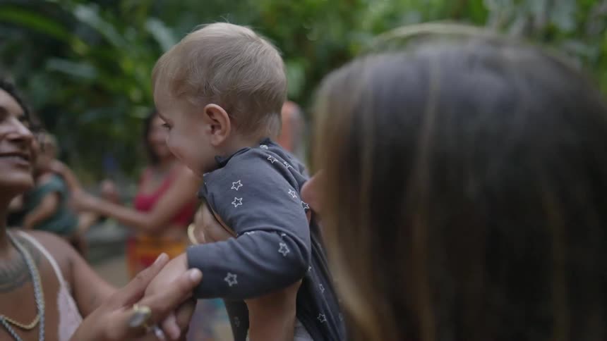 A woman holds up a baby so that another woman can kiss the baby on the cheek as they stand outdoors during the day