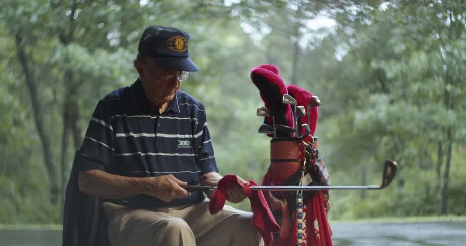 Elderly man wit a dark blue cap polo shirt and beige pants cleans golf club