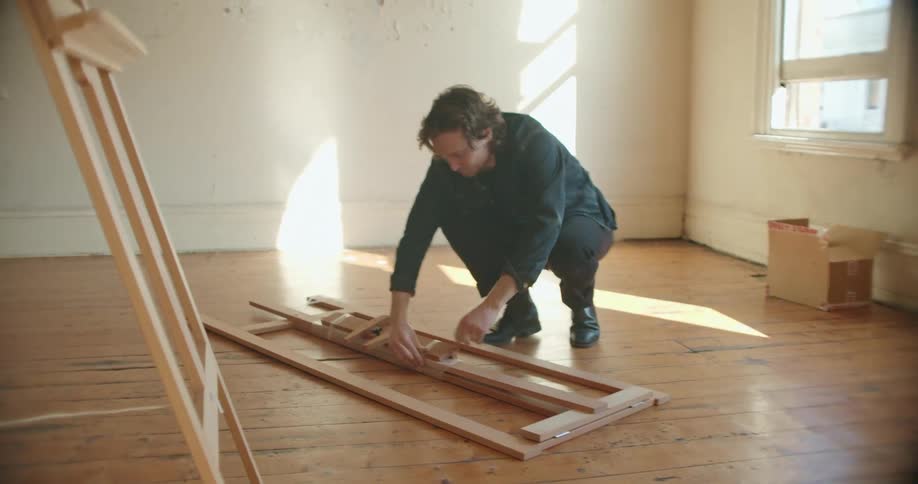 A man in a room untying an easel with a wall and window in the background