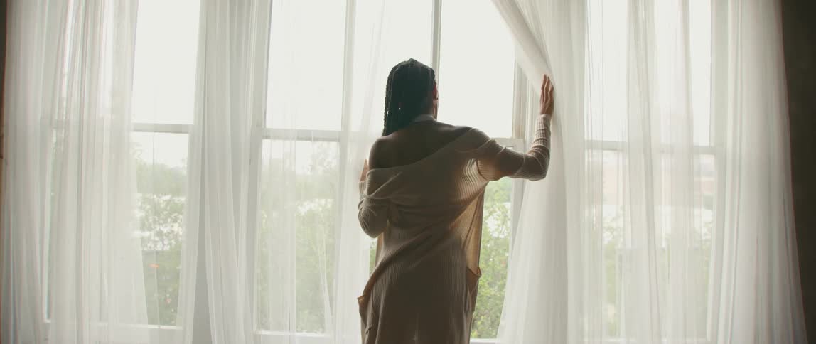 Woman gently moving away the white curtain over the window to look outside