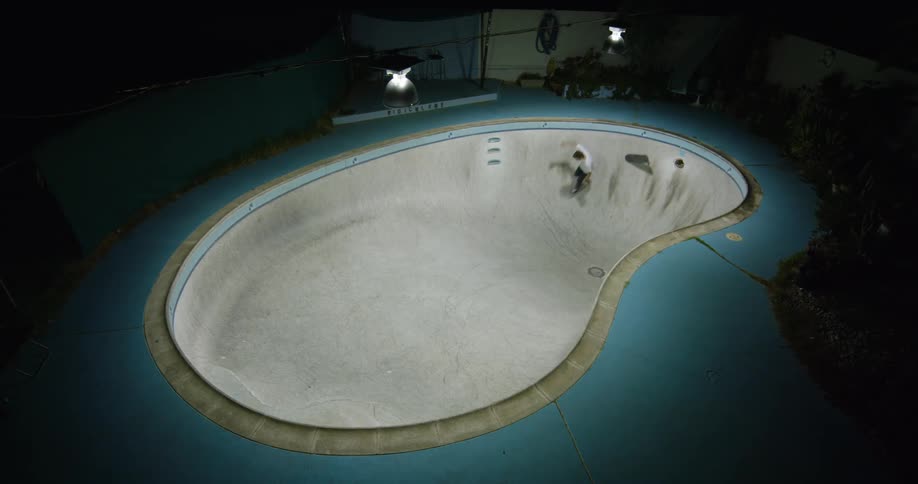 Skateboarder rides skateboard along bottom and side of empty swimming pool in backyard at night