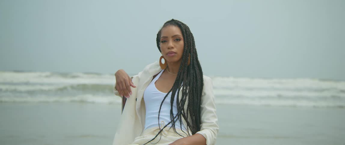 Black woman wearing a large earring sits on the beach near the ocean