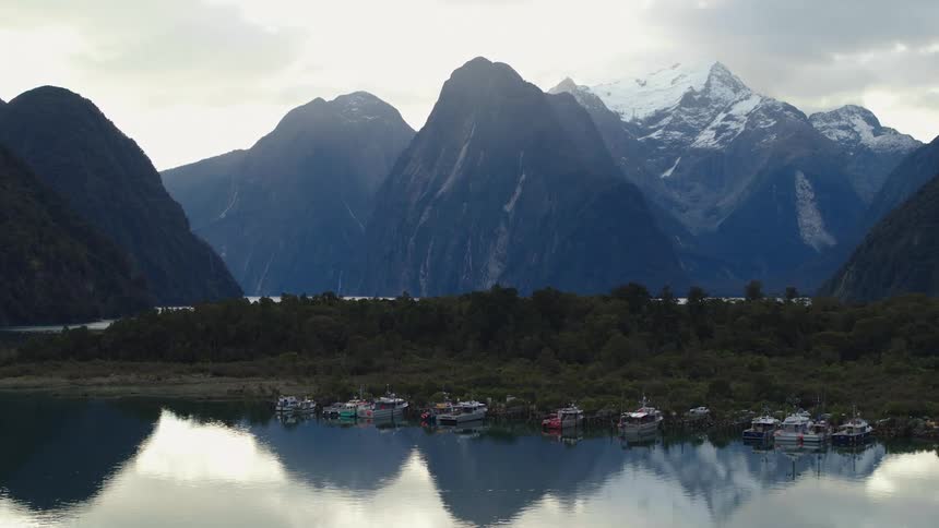 Boats sit near shore on reflective water across wood covered land from more water and snow capped mountains