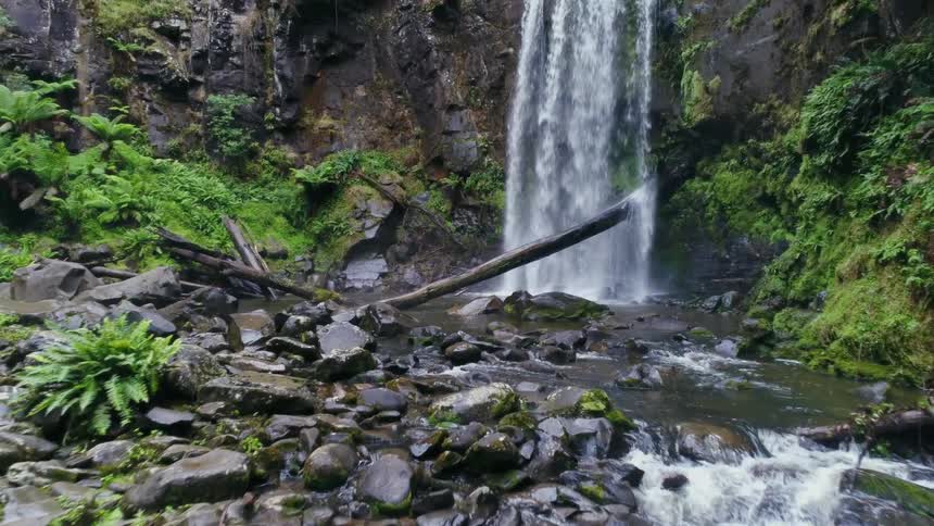 Waterfalls on a steep cliff falls into a stream with rocks and many plants around
