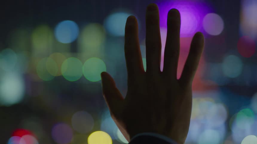 A man holds his hand up in front of lit up lights on buildings in a city