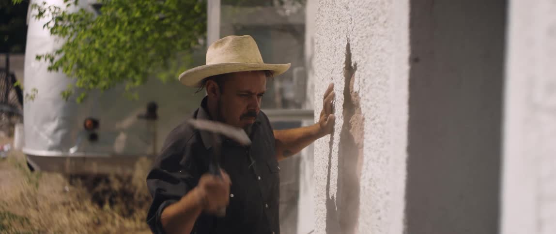 Hispanic man wearing a hat uses a hammer to smash an exterior cement wall of a building
