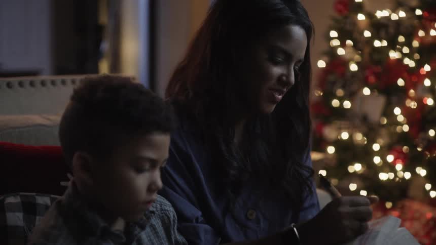 A teenage girl laughs as she sits with her brother next to a lit up christmas tree and laughs