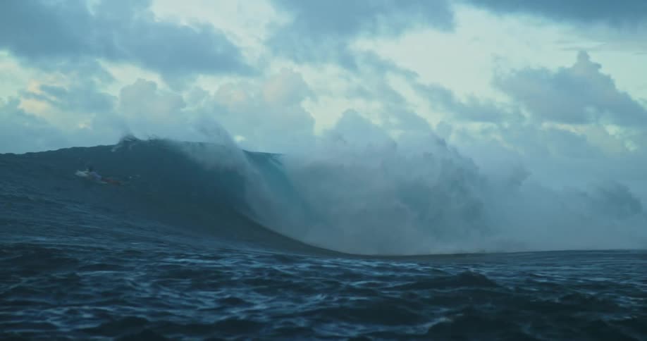 Surfers paddle toward and up a large rolling wave under a cloudy sky