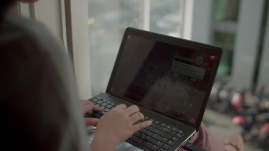 A woman types on her laptop while sitting in front of a large window overlooking a street