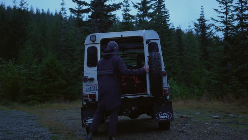 Man walks to land rover parked net to gravel road at edge of conifer forest to access back